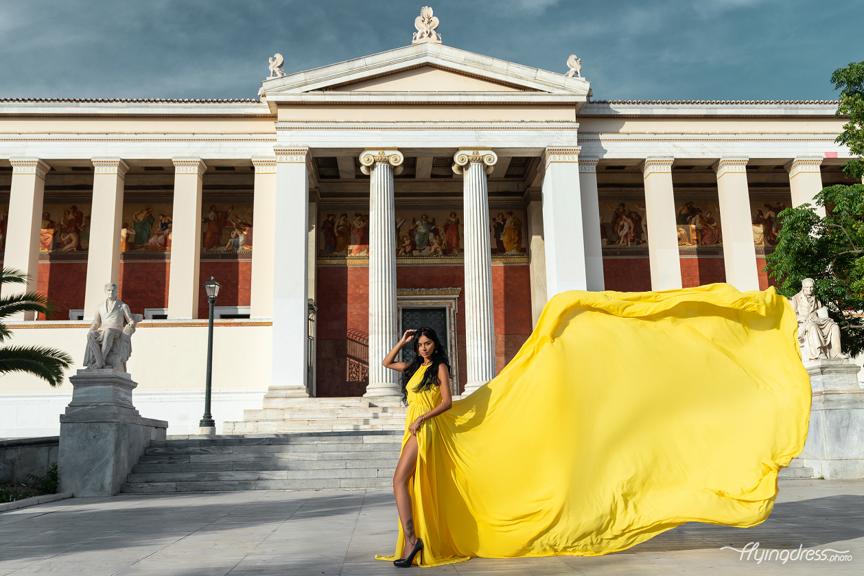 A woman in a flowing yellow dress stands elegantly in front of Athens University, with statues and murals surrounding the grand columns, as the wind lifts the fabric of her dress in a dramatic sweep.